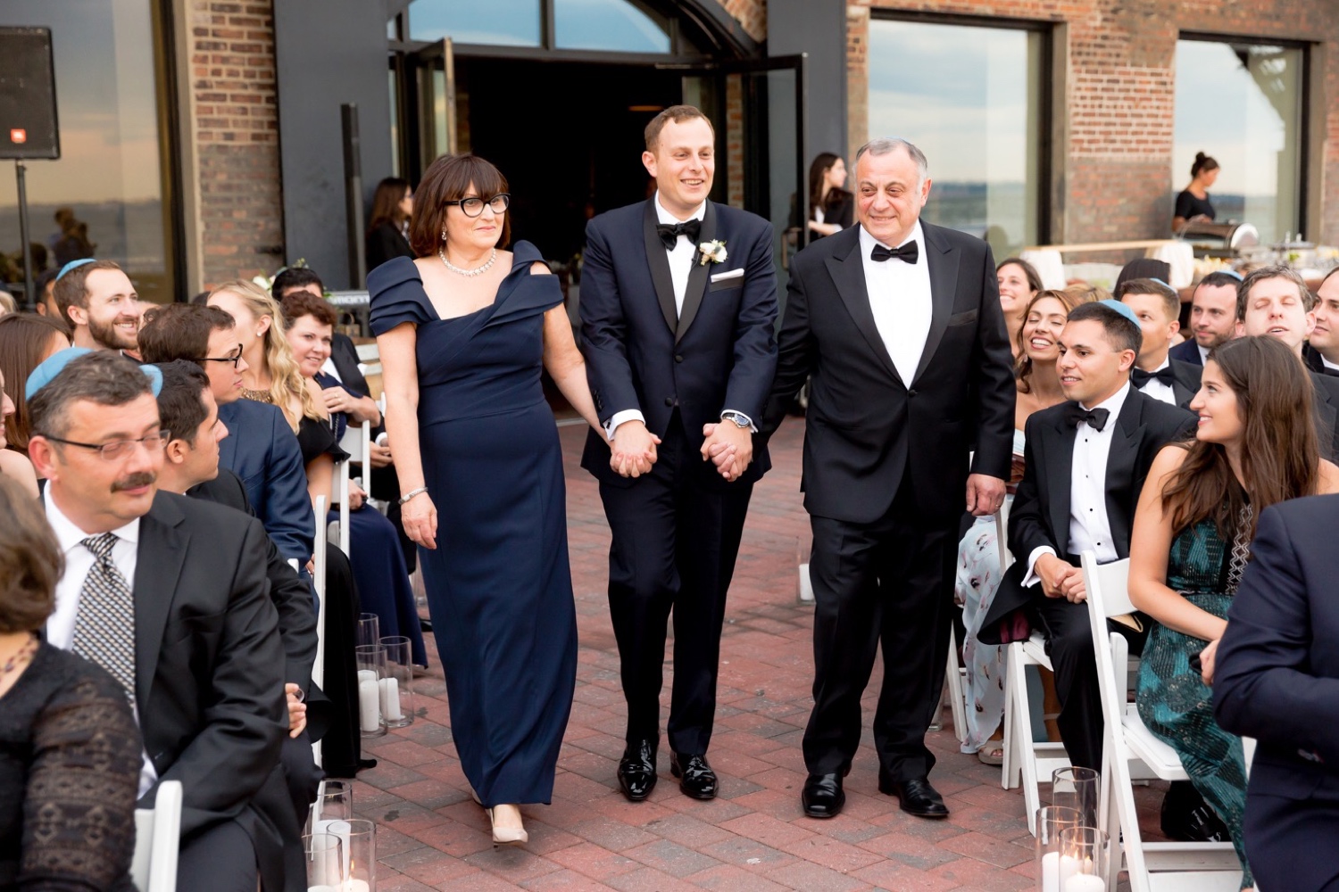 A groom and his parents walking in an aisle during a wedding ceremony at Liberty Warehouse, Brooklyn New York.