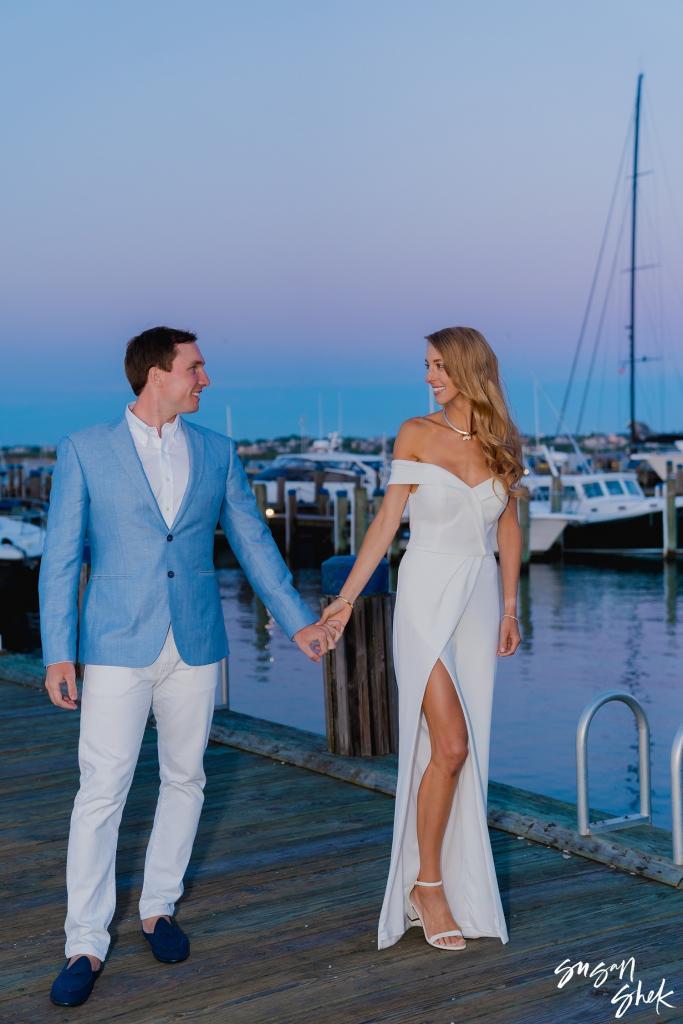 Couple holding hands on a dock in Nantucket harbor at dusk, sailboats behind them