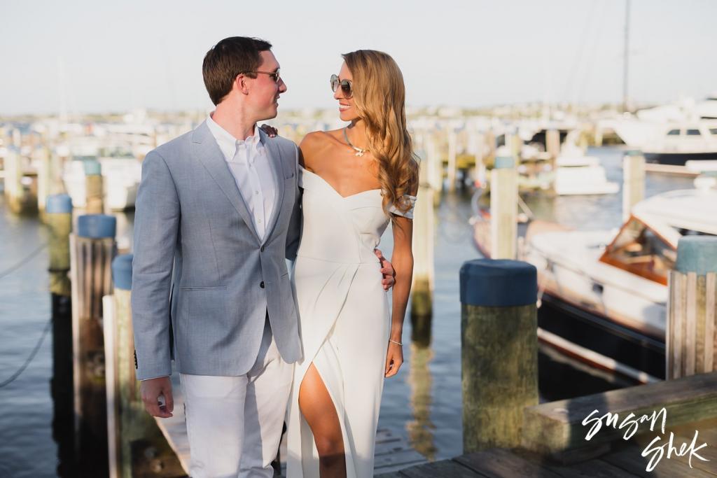 Couple in sunglasses smiling at each other on the docks, boats moored behind them