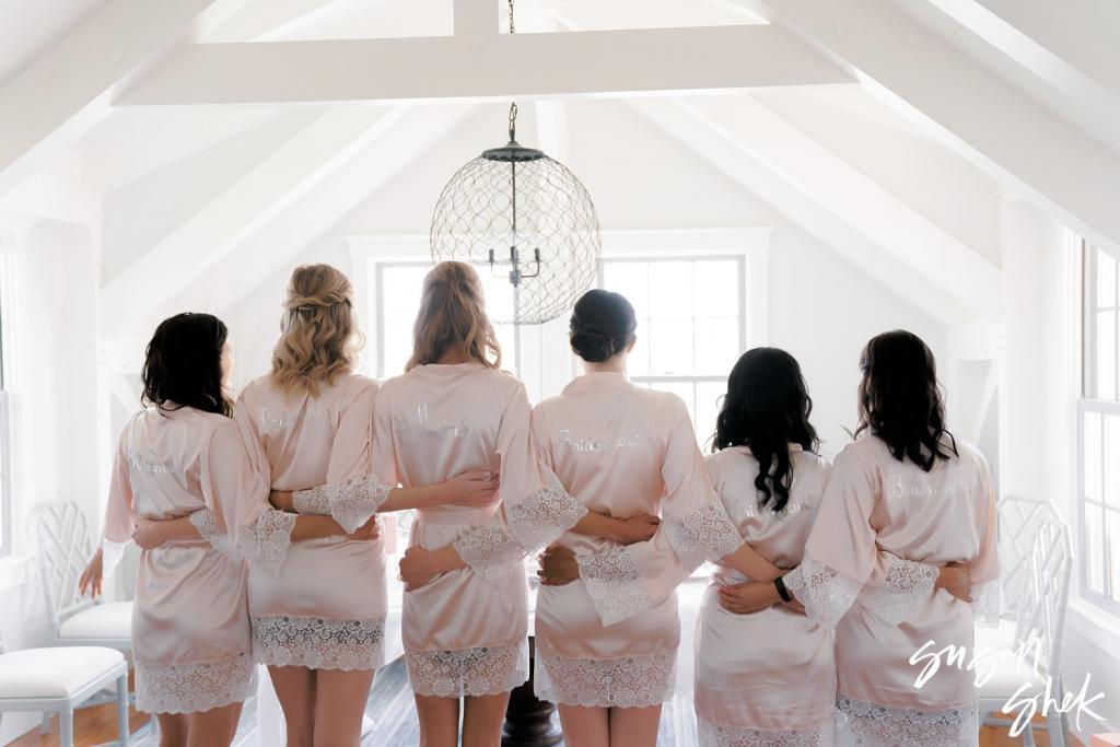 Bride and bridesmaids in blush robes with their arms around each other, seen from behind