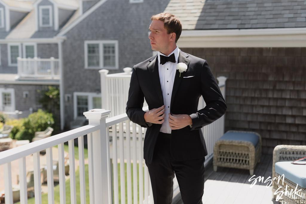Groom fastening his tuxedo jacket on a porch beside shingled houses