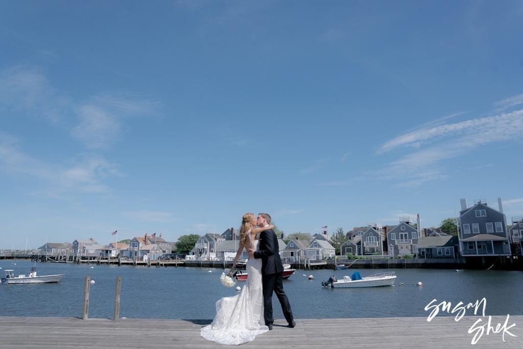 Bride and groom kissing on the dock with the harbor and shingled houses behind them