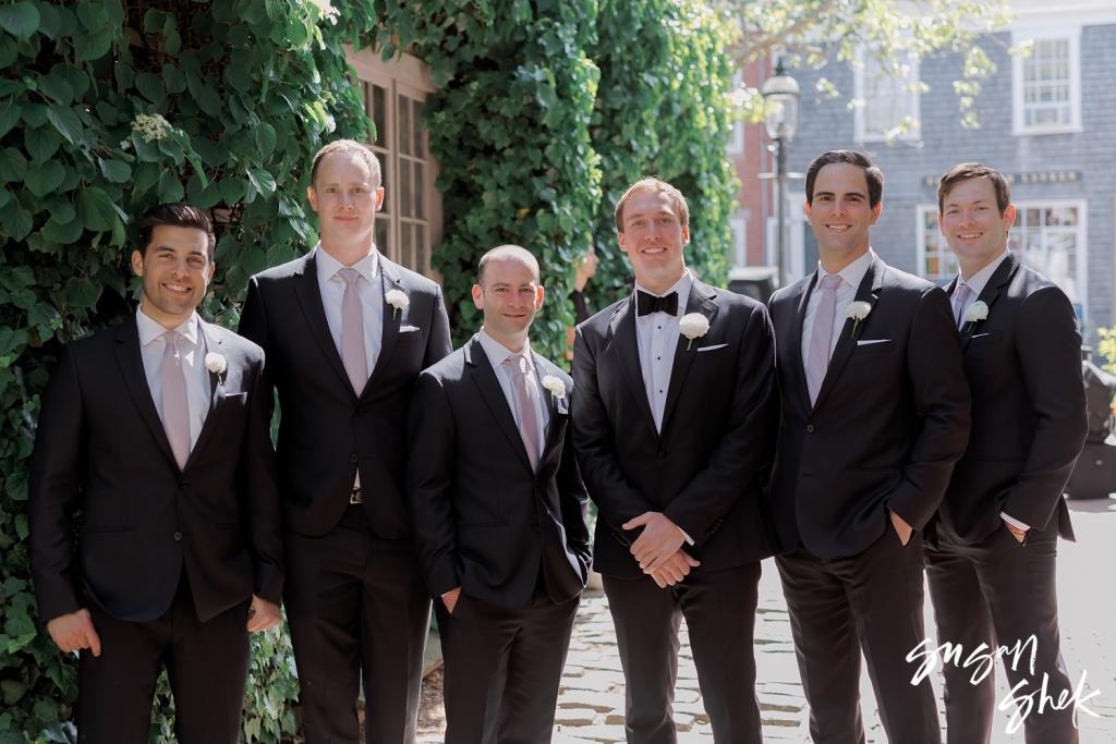 Groomsmen in black tuxedos standing in front of an ivy covered wall