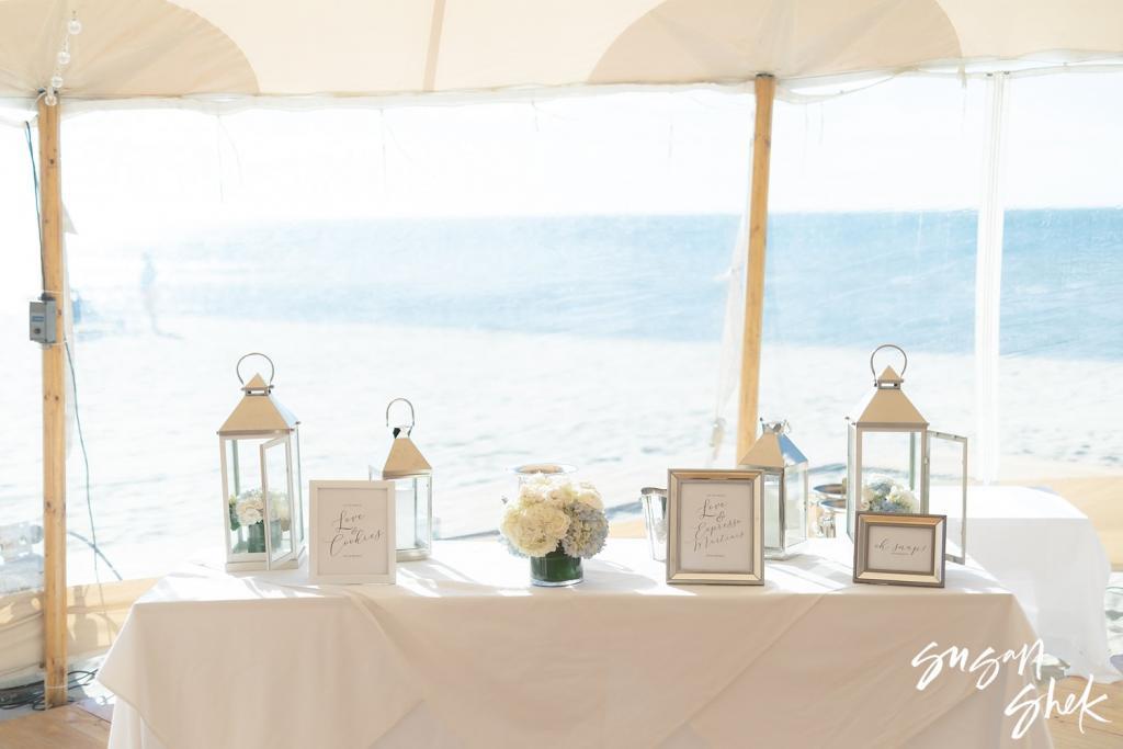 Welcome table with lanterns and framed signs at the edge of the tent, the ocean behind