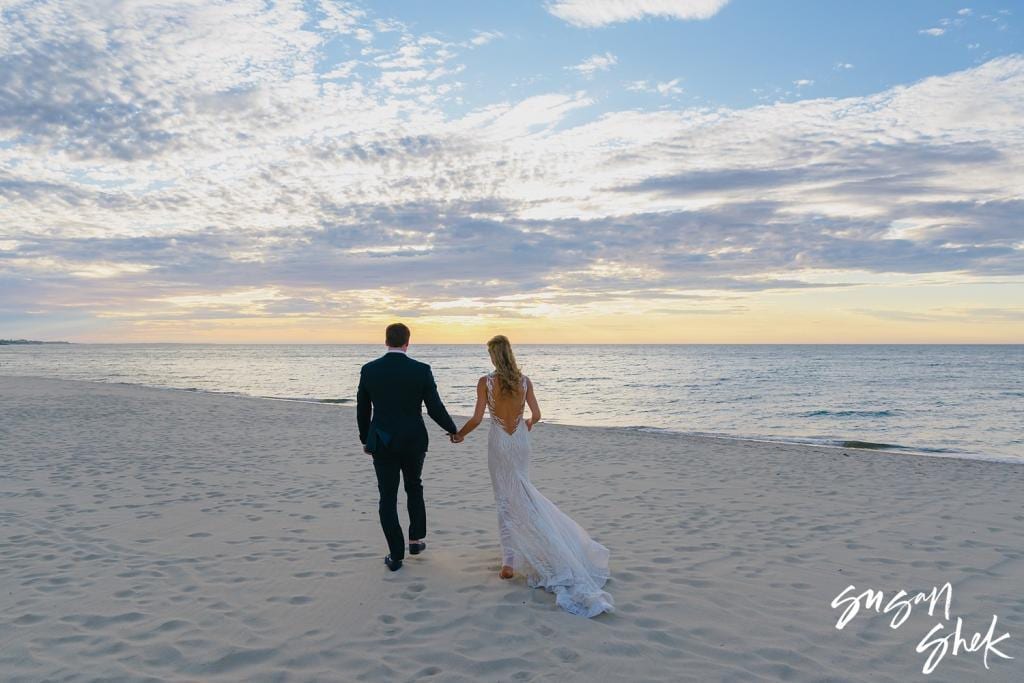 Bride and groom walking hand in hand along the beach toward the water at sunset