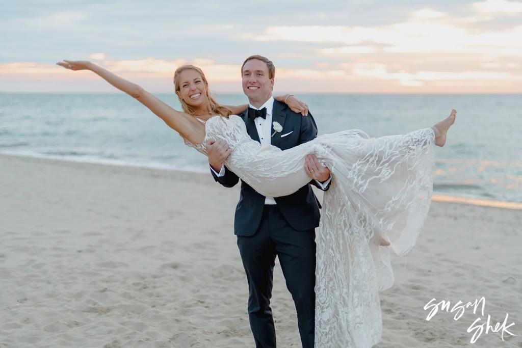 Groom carrying the bride on the beach at sunset as she stretches her arms out