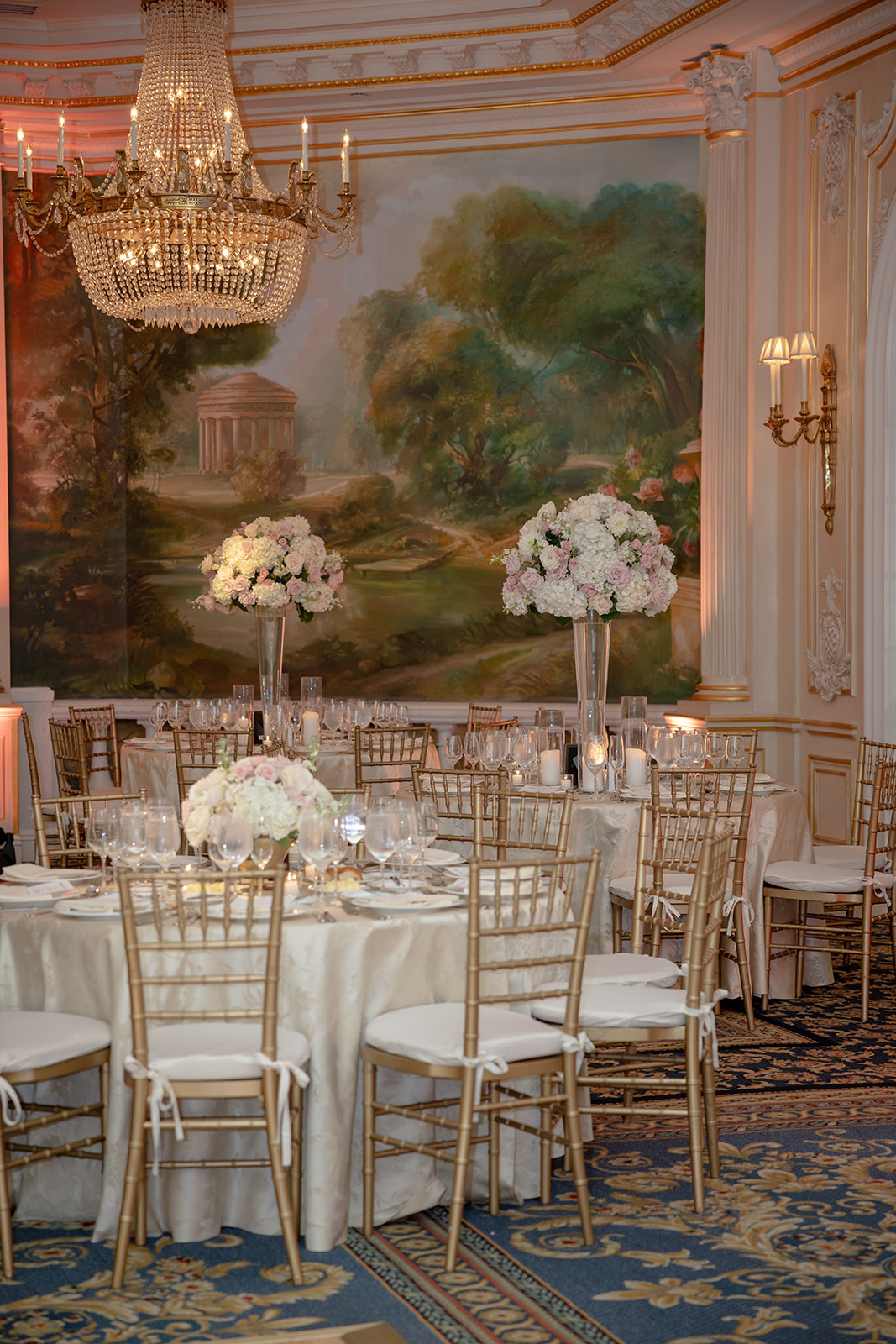 Reception tables arranged with gold seating, candlelight, and towering floral arrangements beside painted mural walls.