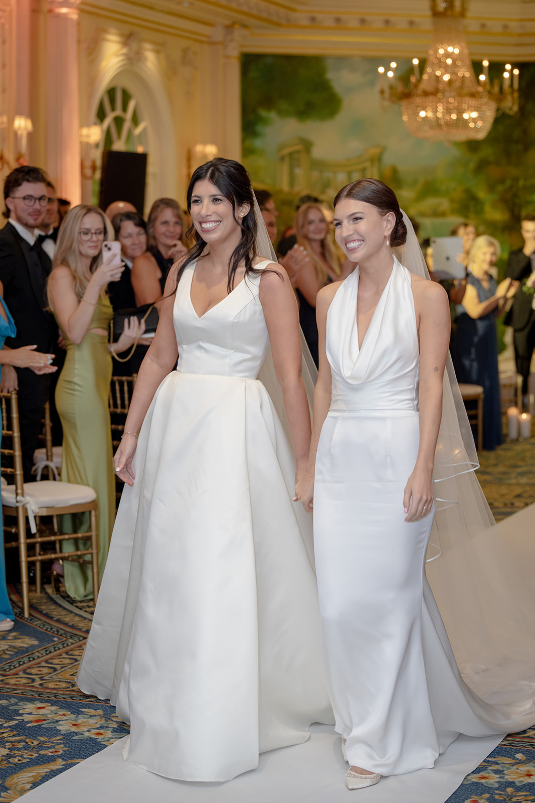 Two brides walking down the ceremony aisle hand in hand at their Essex House NYC wedding while guests cheer.
