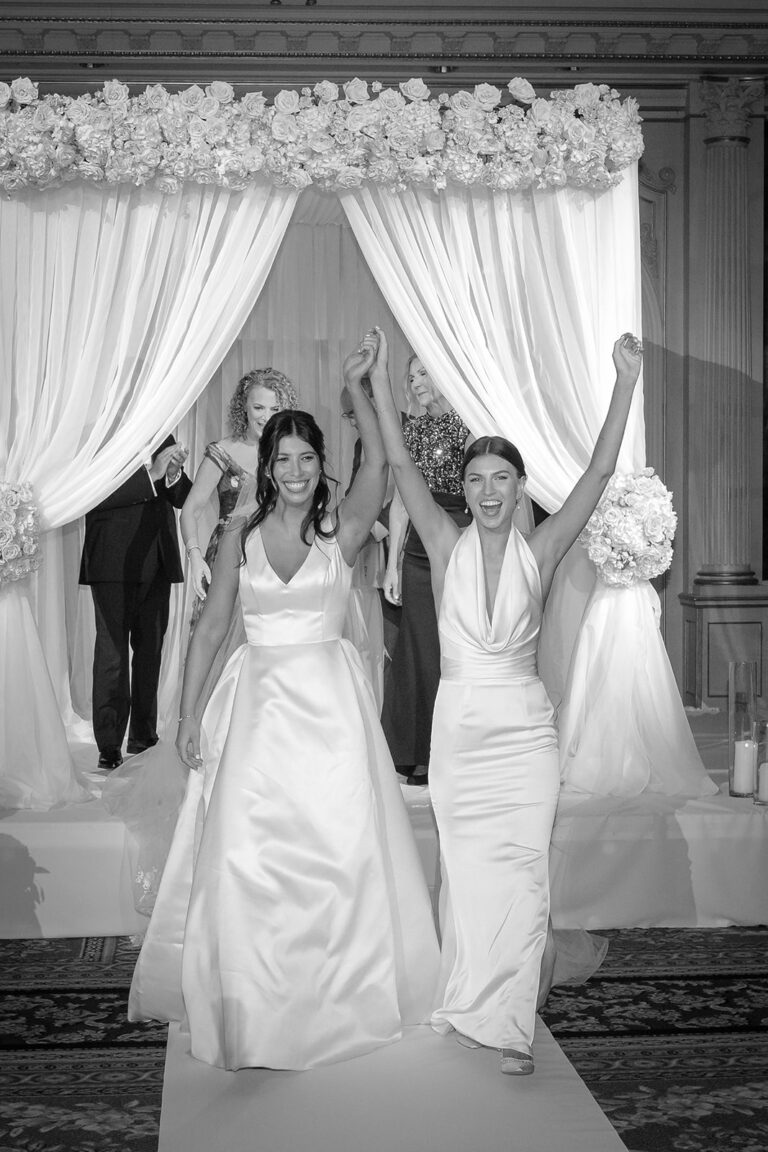 Black-and-white photo of two brides celebrating beneath their floral chuppah at the JW Marriott Essex House NYC wedding ceremony.
