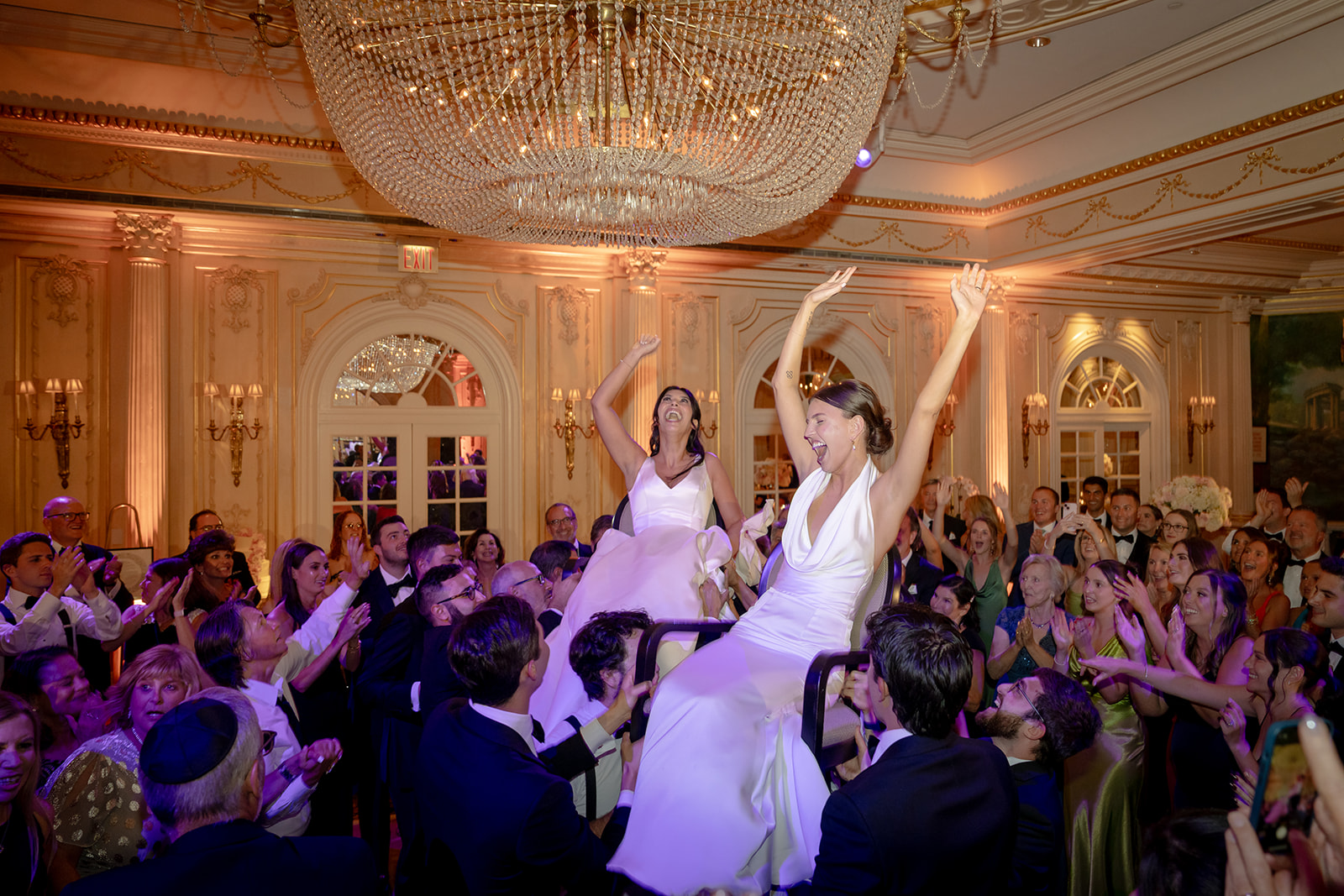 The brides lifted during the hora celebration, surrounded by cheering guests beneath a grand chandelier.