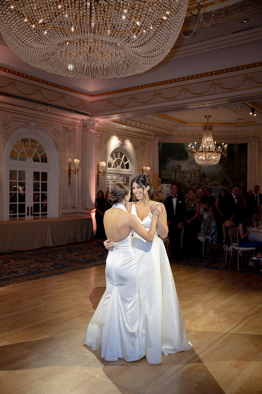 Romantic first dance between the brides under sparkling chandelier lighting.