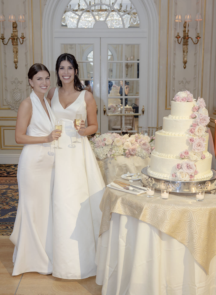 Two brides cheering with champagne glasses next to their  cake at the Essex House wedding 