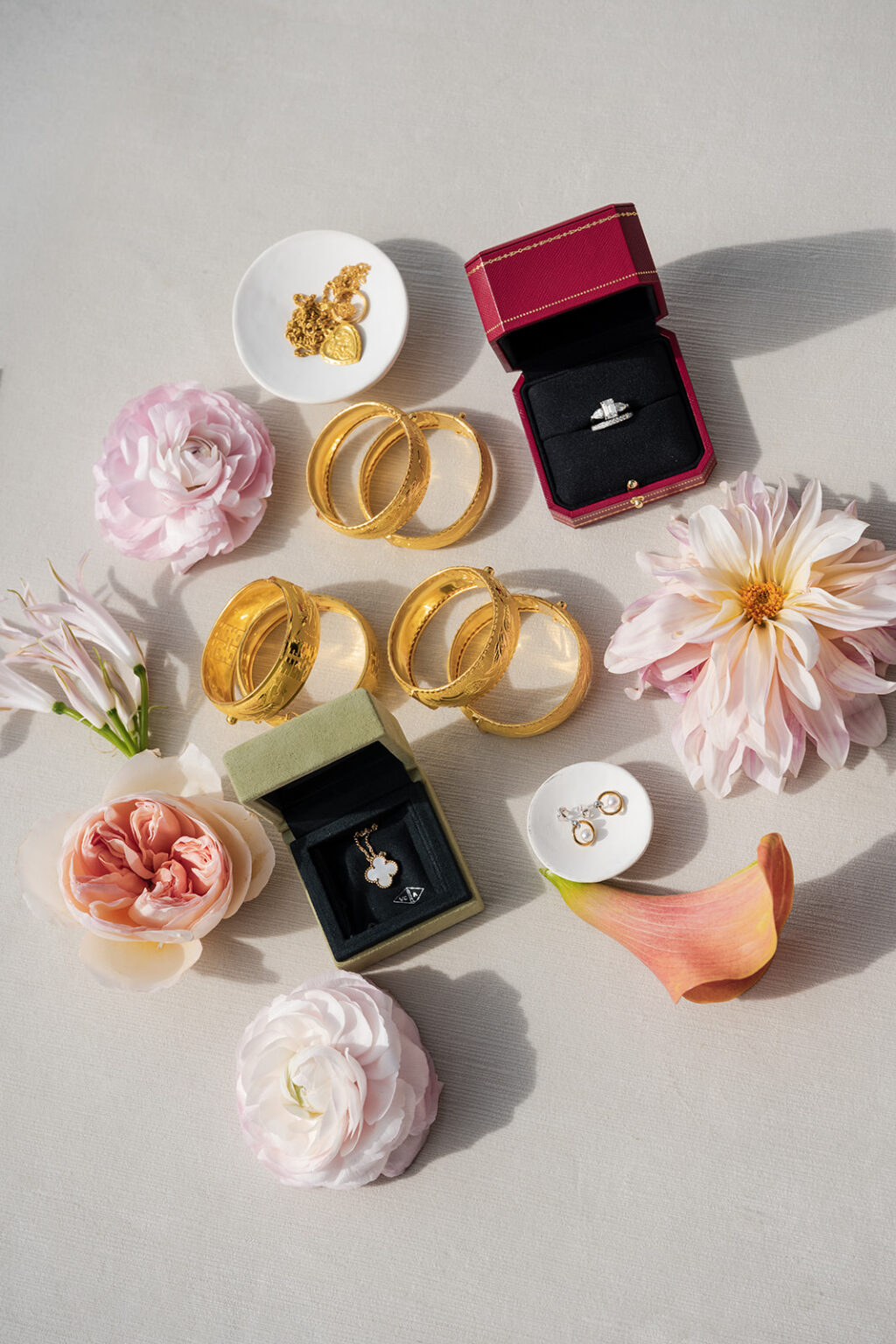 Traditional gold wedding jewelry and engagement ring styled in red ring boxes with soft pastel flowers photographed in clean editorial lighting.