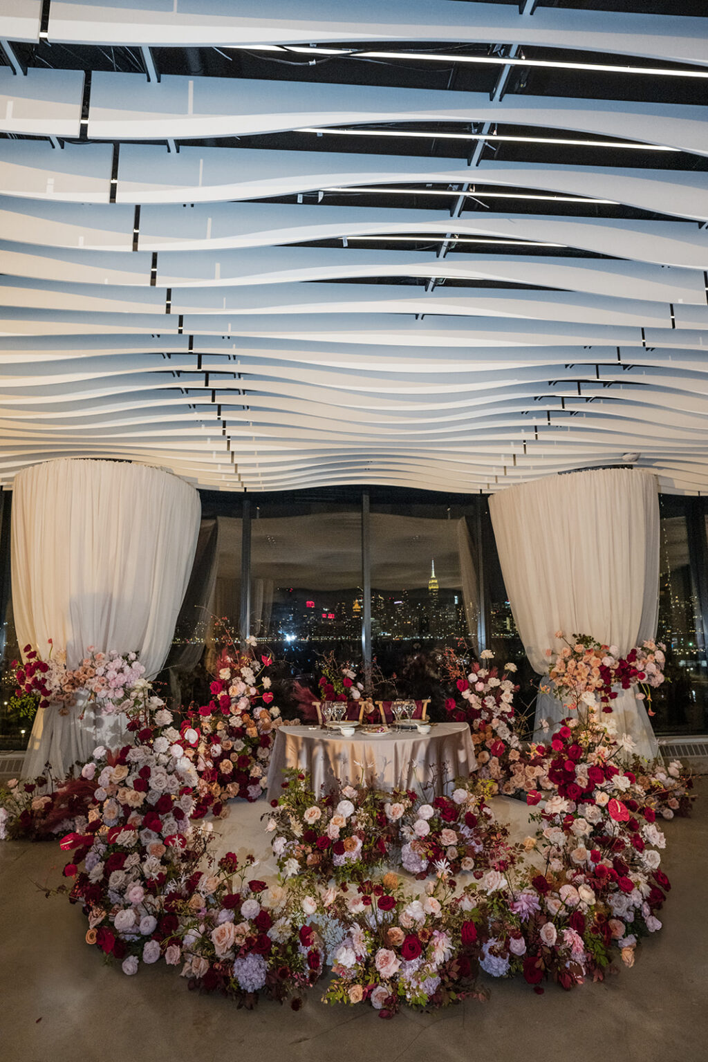 Romantic sweetheart table at Vesper Events with dramatic layered florals and city skyline visible through floor to ceiling windows at night.