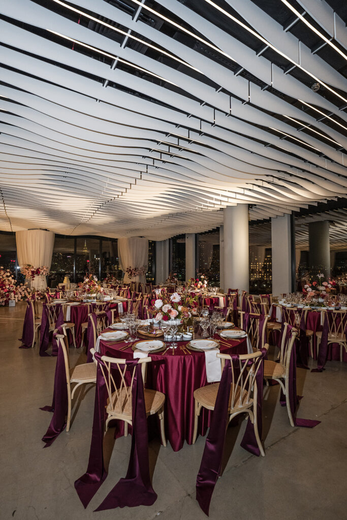 Wide reception view with burgundy table linens, gold chargers, floral centerpieces, and sculptural ceiling design illuminated for evening celebration.