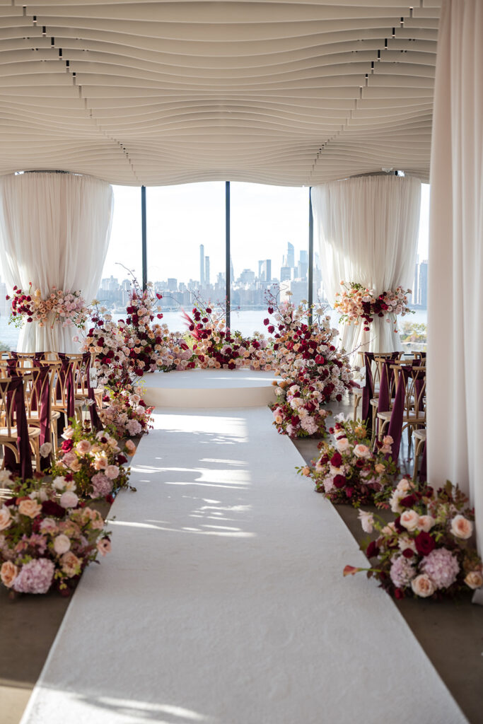 Ceremony setup at Vesper Events with a floral lined aisle, layered pink and burgundy arrangements, and floor to ceiling windows overlooking the NYC skyline.