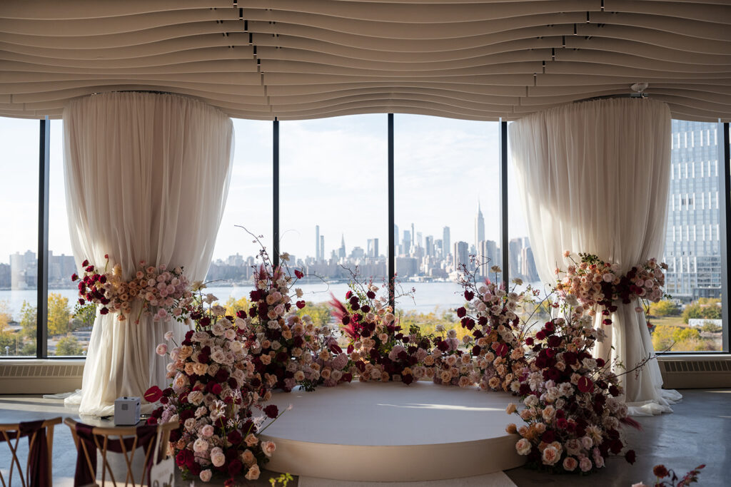 Modern waterfront ceremony design featuring abundant roses, textured florals, soft draping, and city views through tall glass windows.