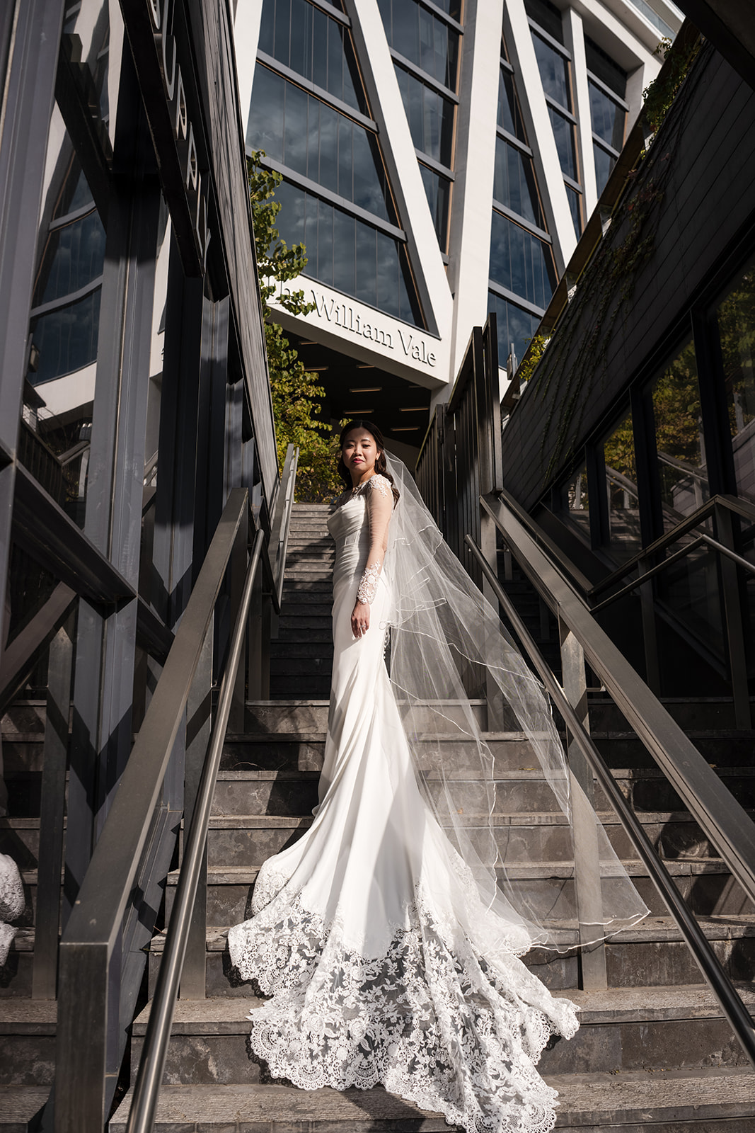 Bride standing on modern stone stairs outside The William Vale hotel with long lace train flowing dramatically in natural sunlight.
