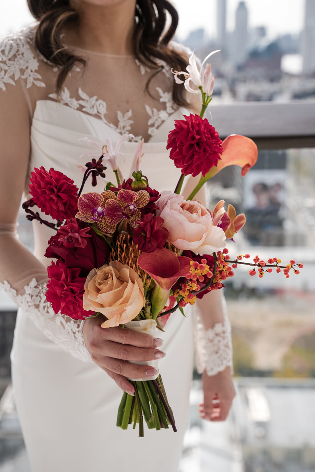 Close up of bridal bouquet featuring deep red dahlias, blush roses, orchids, and calla lilies photographed in soft natural light.