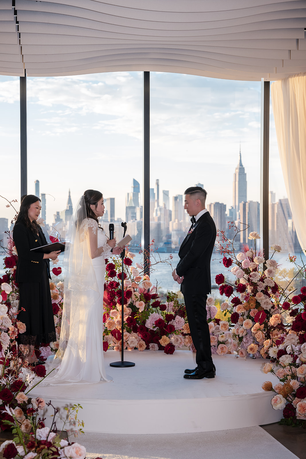 Wedding ceremony at Vesper Events overlooking the NYC skyline as the couple exchange vows surrounded by abundant pink and burgundy florals.