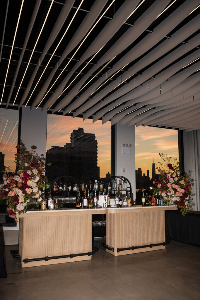 Modern wedding bar setup at sunset with skyline views, curated spirits display, and dramatic evening lighting.