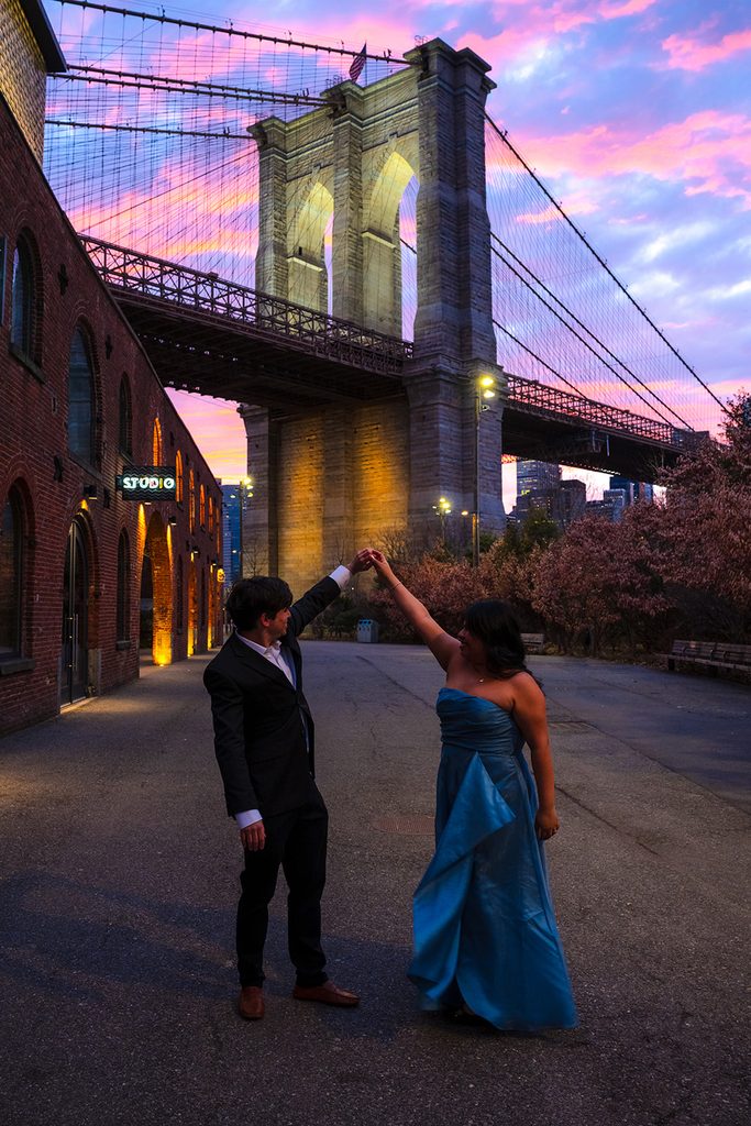 Couple dancing on a cobblestone street beneath the Brooklyn Bridge at dusk