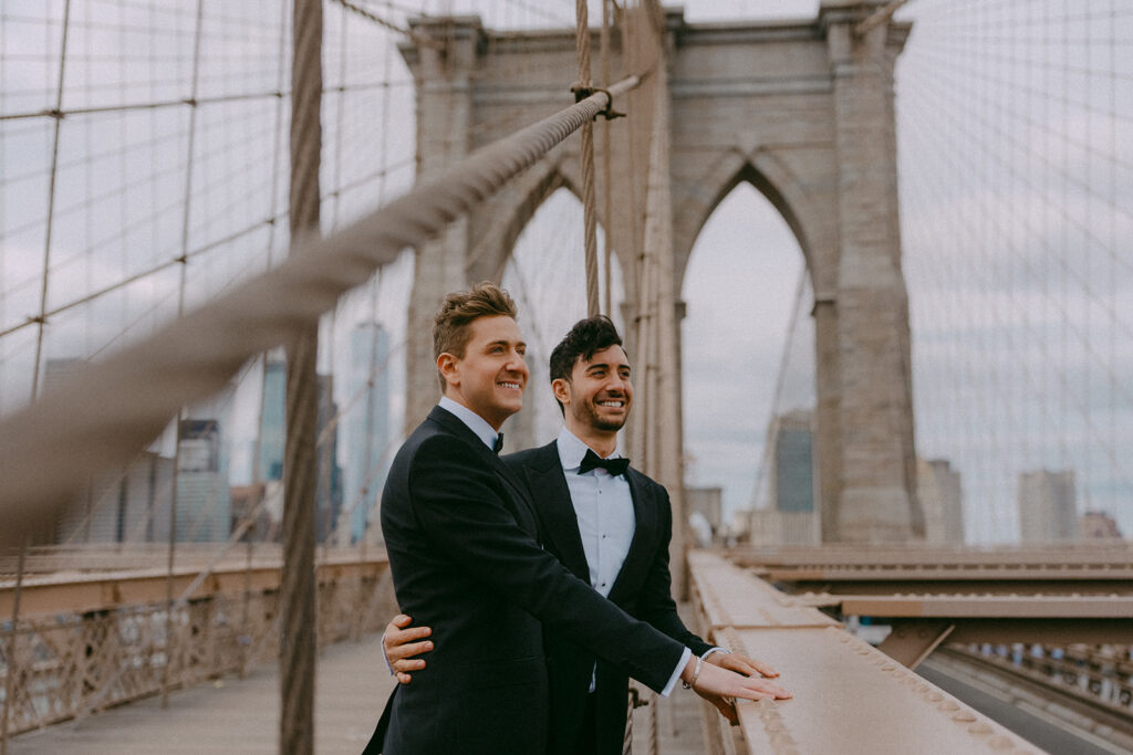 Two grooms laughing on the Brooklyn Bridge with the Manhattan skyline behind them