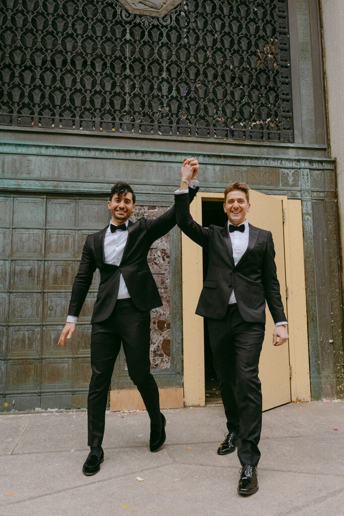 Two grooms cheering as they walk out through bronze doors after the ceremony
