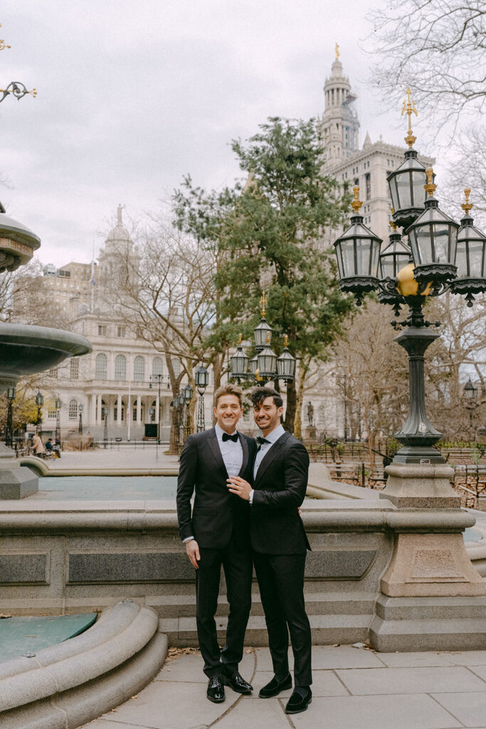 Two grooms in tuxedos by a fountain and old lamp posts in a Lower Manhattan park