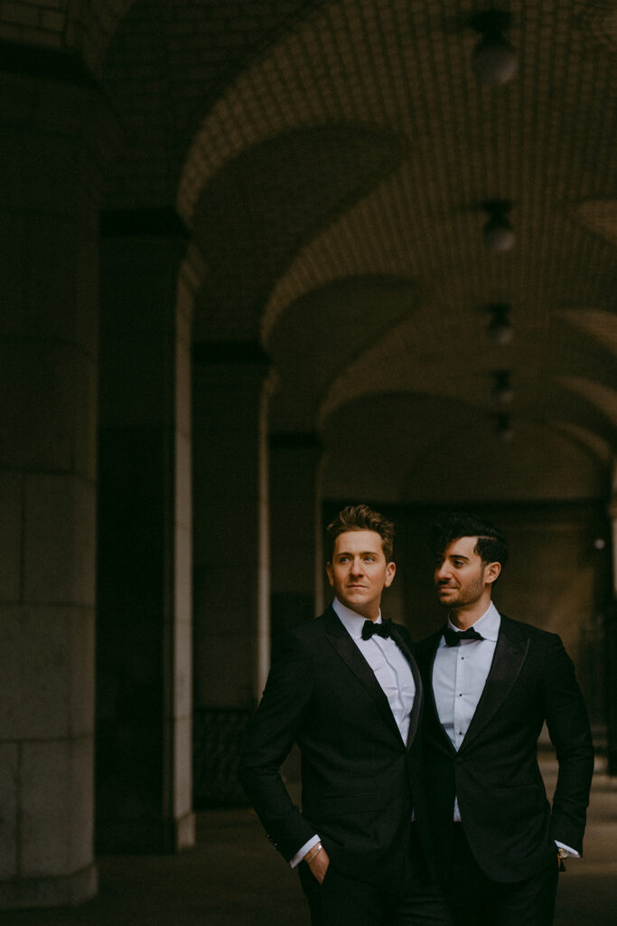 Two grooms in tuxedos standing in a dim vaulted arcade