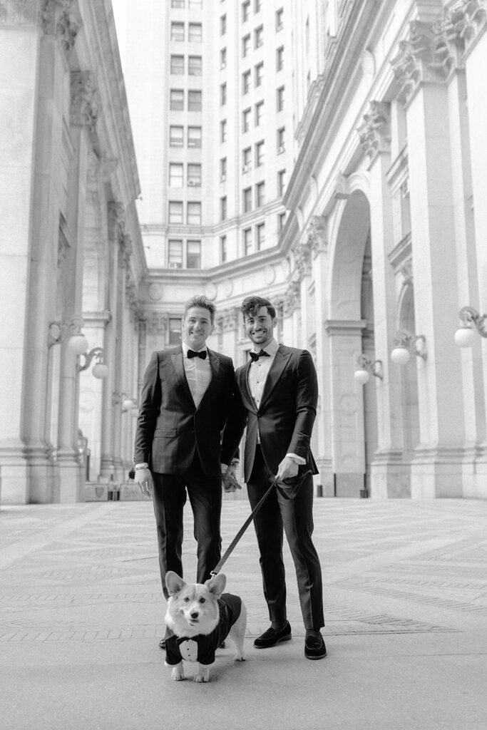Black and white photo of two grooms walking their corgi past columned buildings